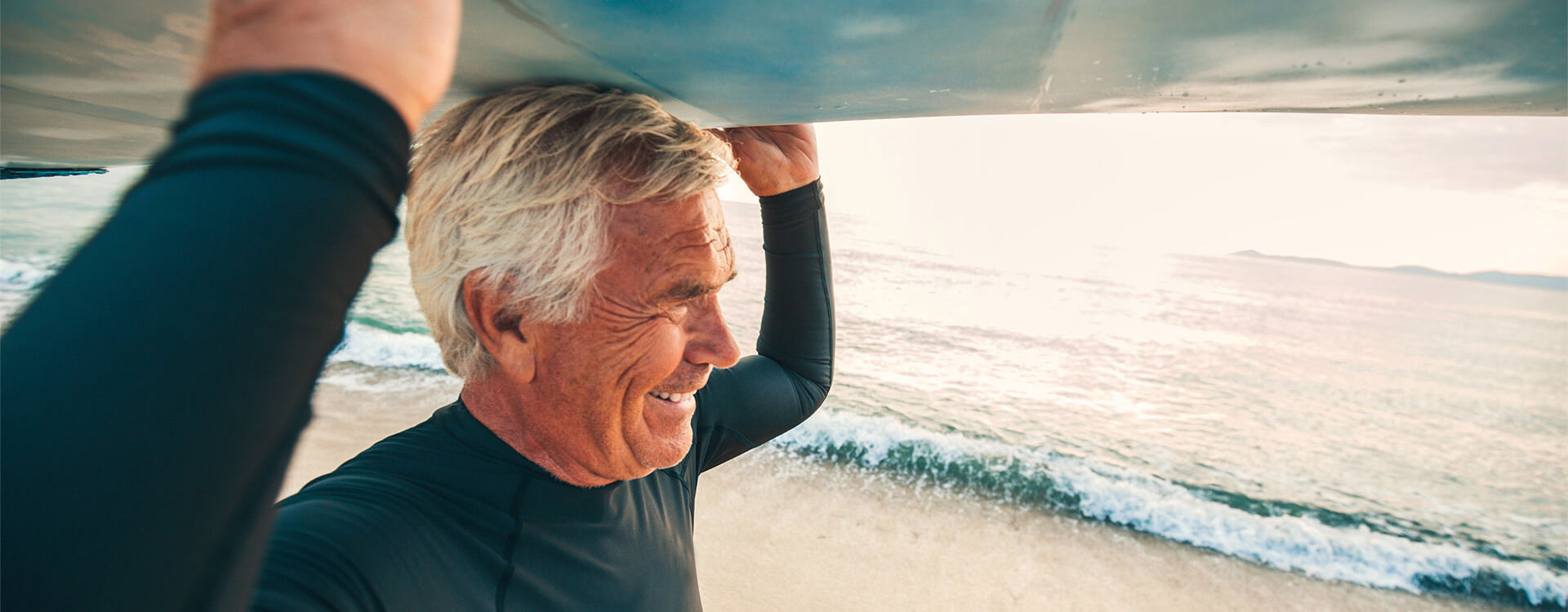 Älterer Mann im Neoprenanzug trägt ein Surfbrett zum Meer