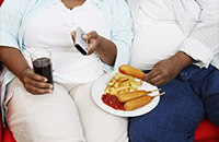 Zwei beleibte Menschen sitzen auf dem Sofa und essen fettiges Essen, trinken Cola und sehen fern.
