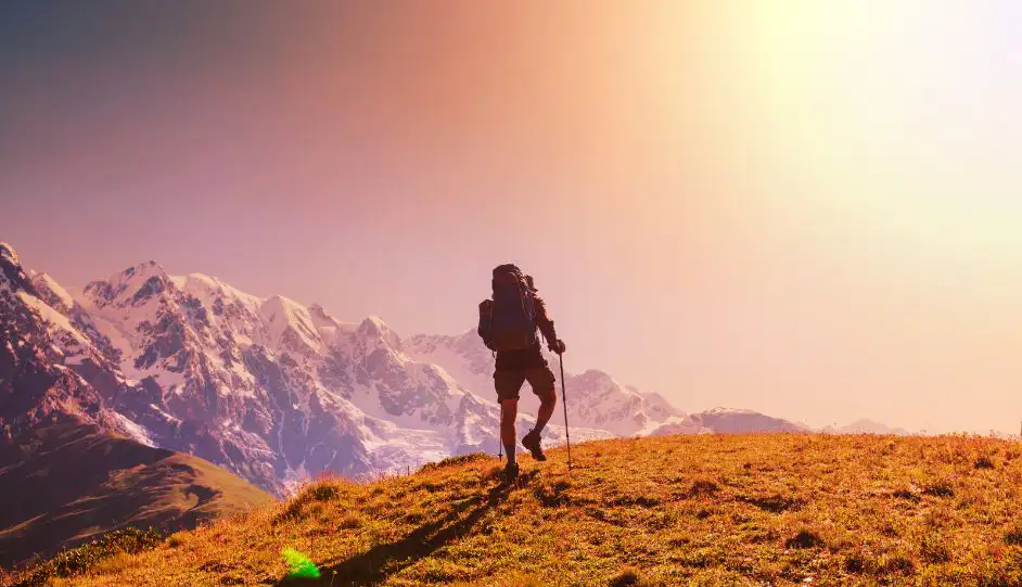 Wanderer mit Rucksack und Wanderstock auf Berggipfel bei Sonnenuntergang, schneebedeckte Alpen im Hintergrund